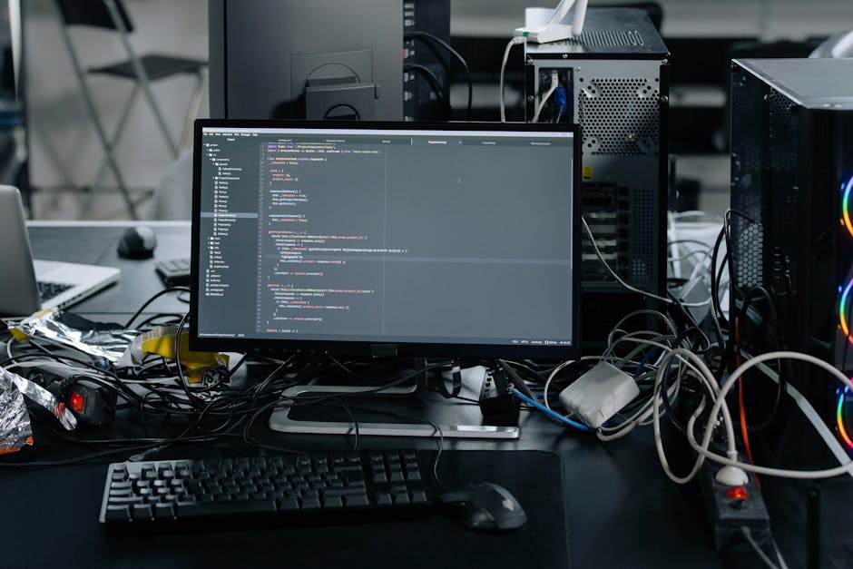 A cluttered desk setup featuring a central computer monitor displaying a coding interface with syntax highlighted code in different colours. Surrounding the monitor are various tangled cables, includi