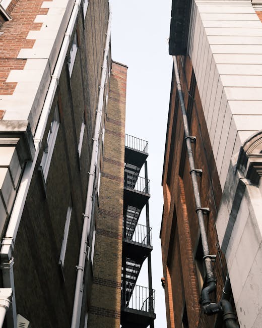 An upward view between two closely situated buildings on Shoreditch High Street, showing a narrow passageway with a black metal fire escape staircase attached to the brick wall on the left side. The b